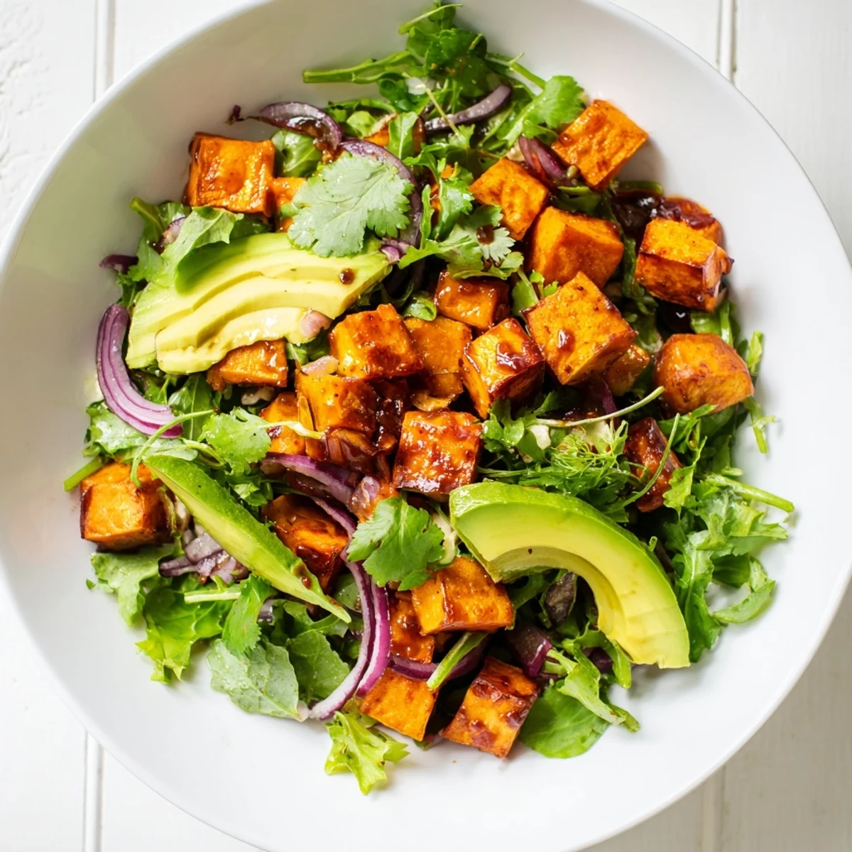 A vibrant vegetarian roasted sweet potato salad with avocado slices, mixed greens, and a lime dressing, served with lime wedges on the side.