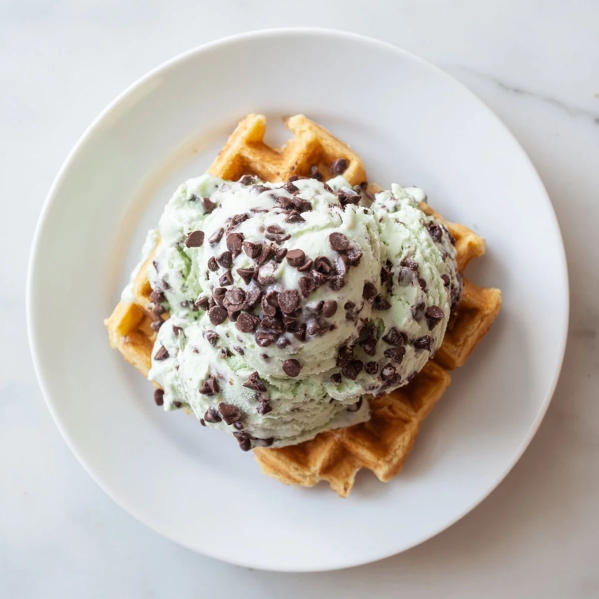 A warm, golden waffle topped with scoops of homemade mint chocolate chip ice cream, showing creamy green swirls and chocolate flecks.