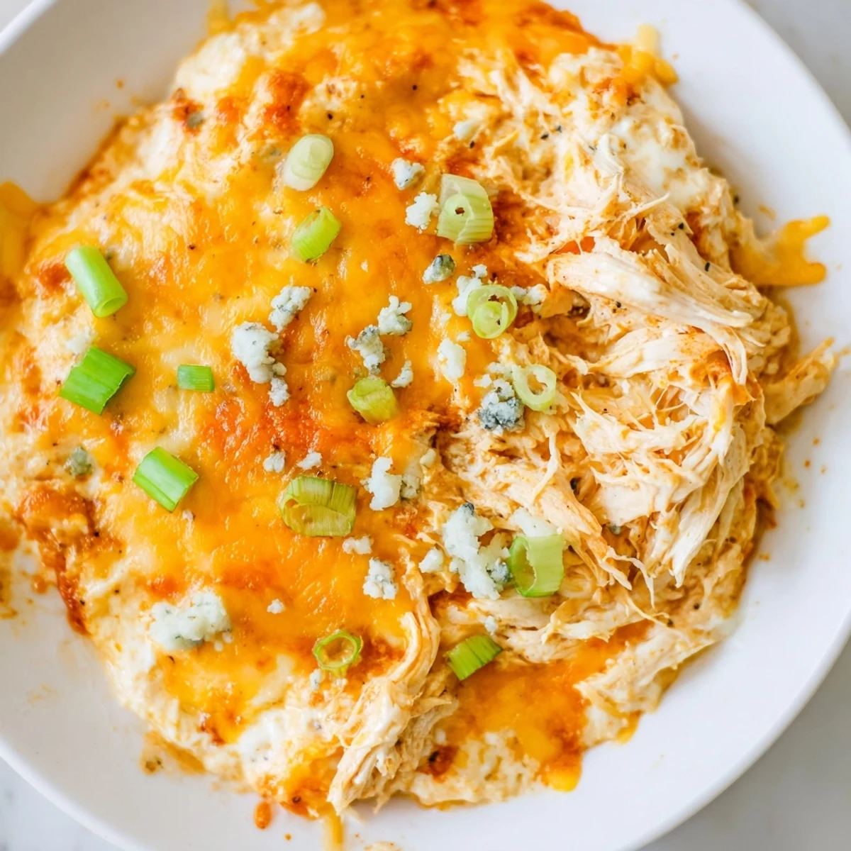 Golden-baked Keto Buffalo Chicken Dip in a white dish, topped with melted mozzarella and cheddar cheese, garnished with fresh green onions and crumbled blue cheese.
