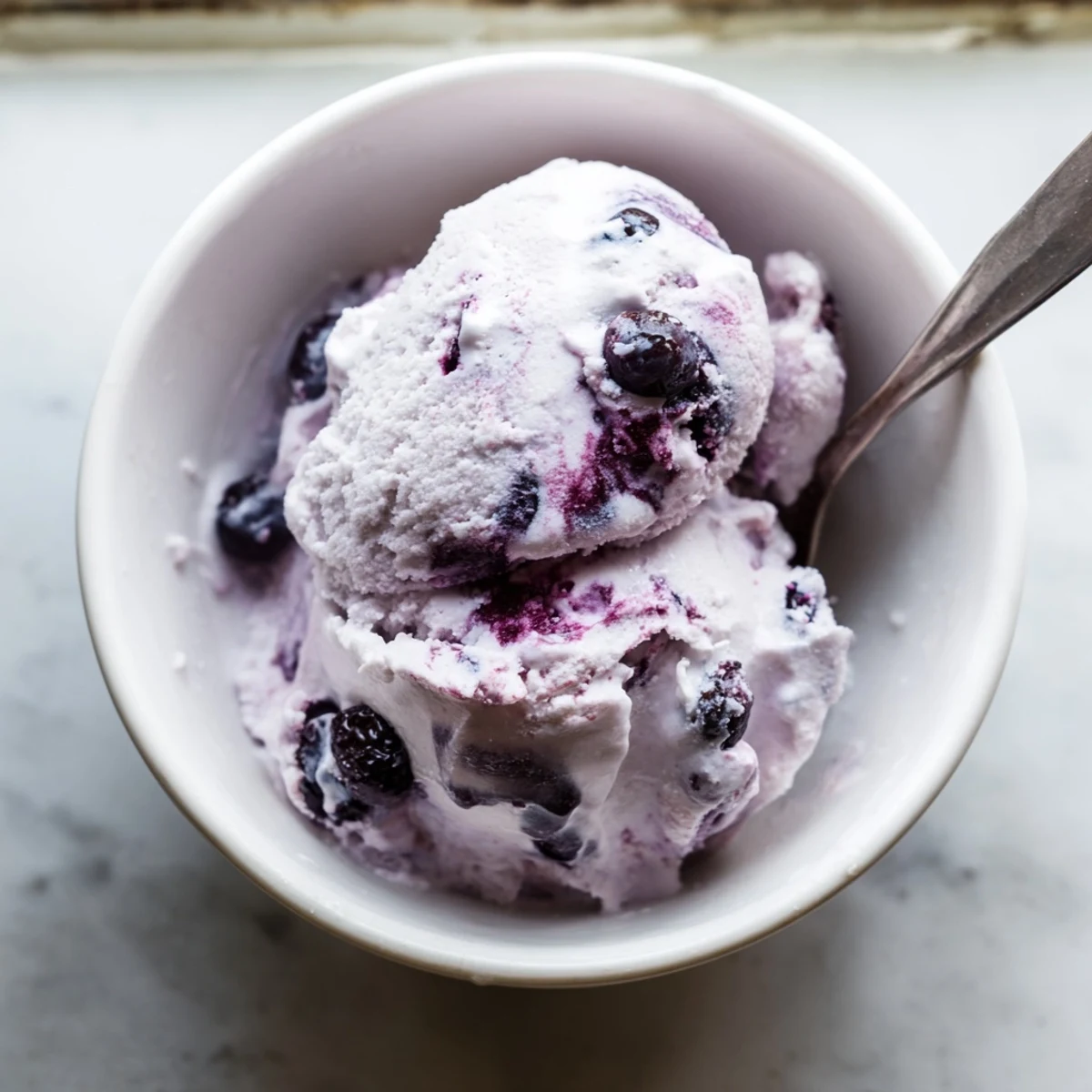 A close-up of creamy Blueberry Cottage Cheese Ice Cream scoops glistening with honey glaze, fresh blueberries, and a mint garnish in a rustic white bowl.