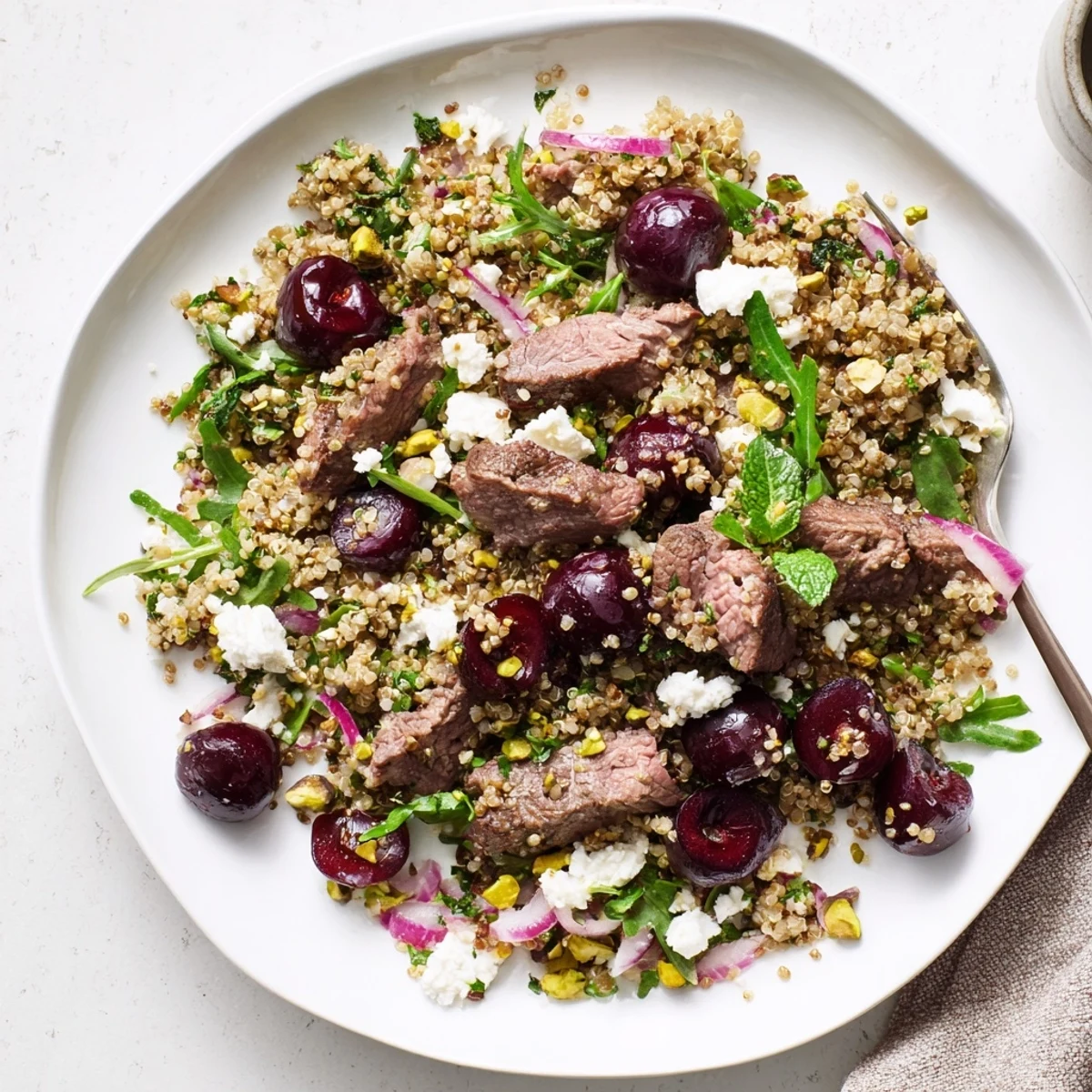 Mediterranean-style cherry quinoa salad with lamb plated with toasted pistachios sweet halved cherries and sliced grilled lamb on a bed of greens