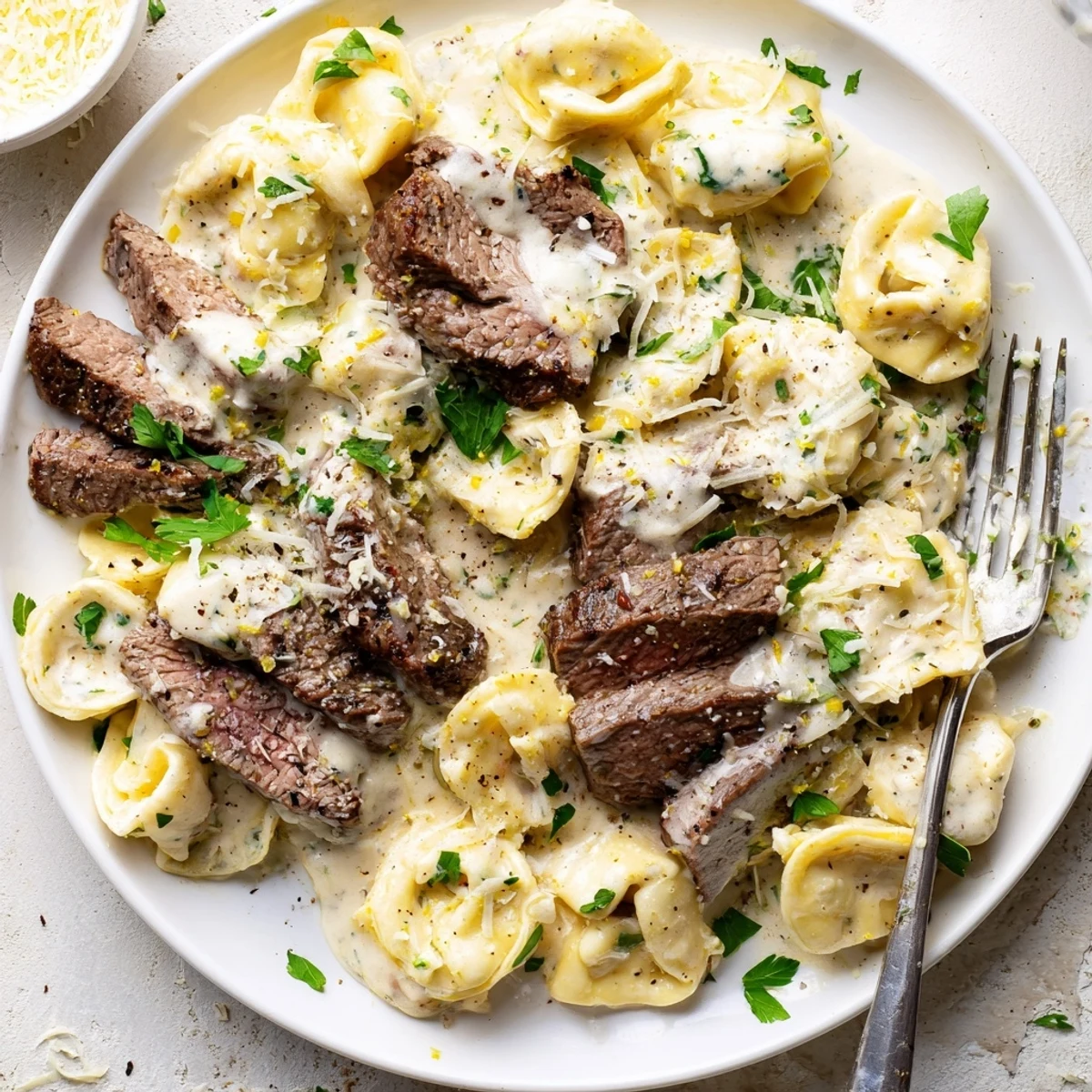 Creamy garlic steak tortellini dish topped with fresh Parmesan and lemon zest in white serving bowl